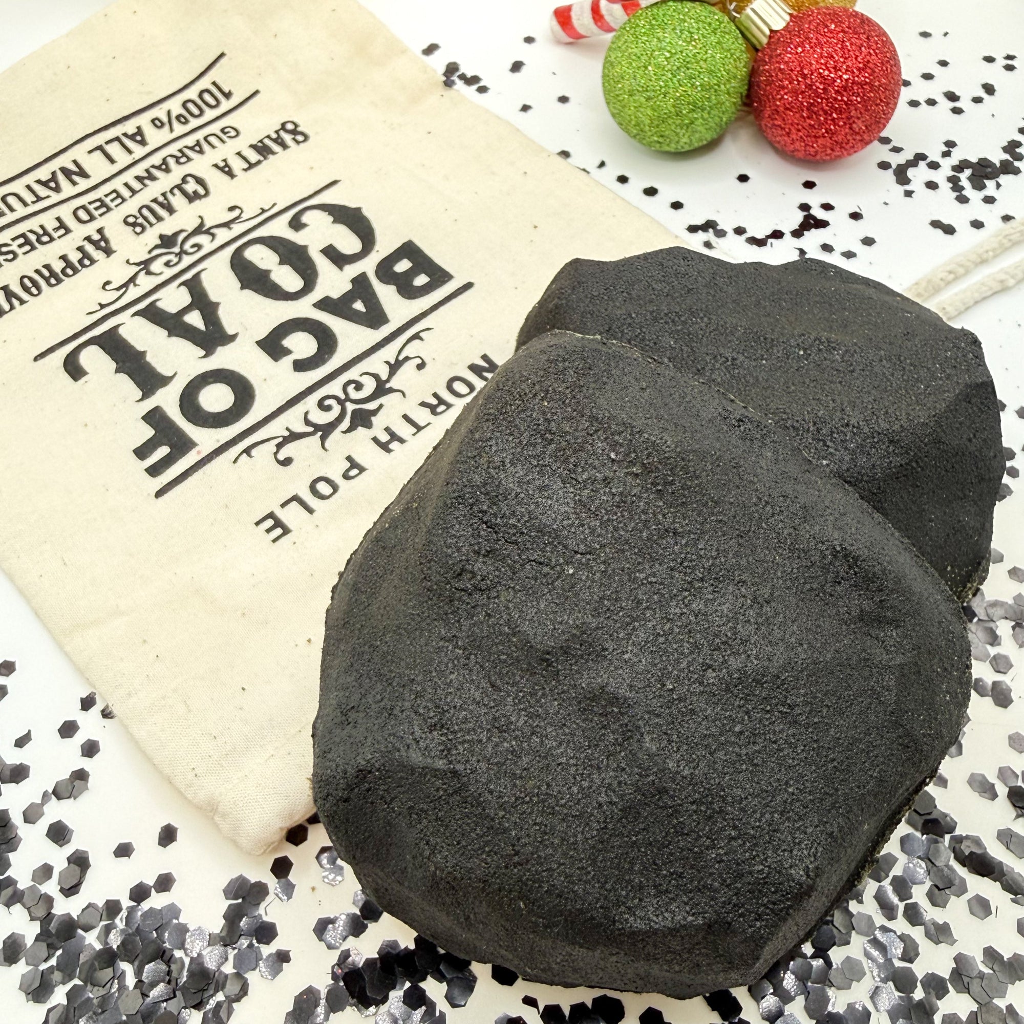 Two black lump of coal bath bomb gag gift with a tan canvas bag labeled 'Bag of Coal' on a decorative surface handmade by Rusted Acre Soap Co.