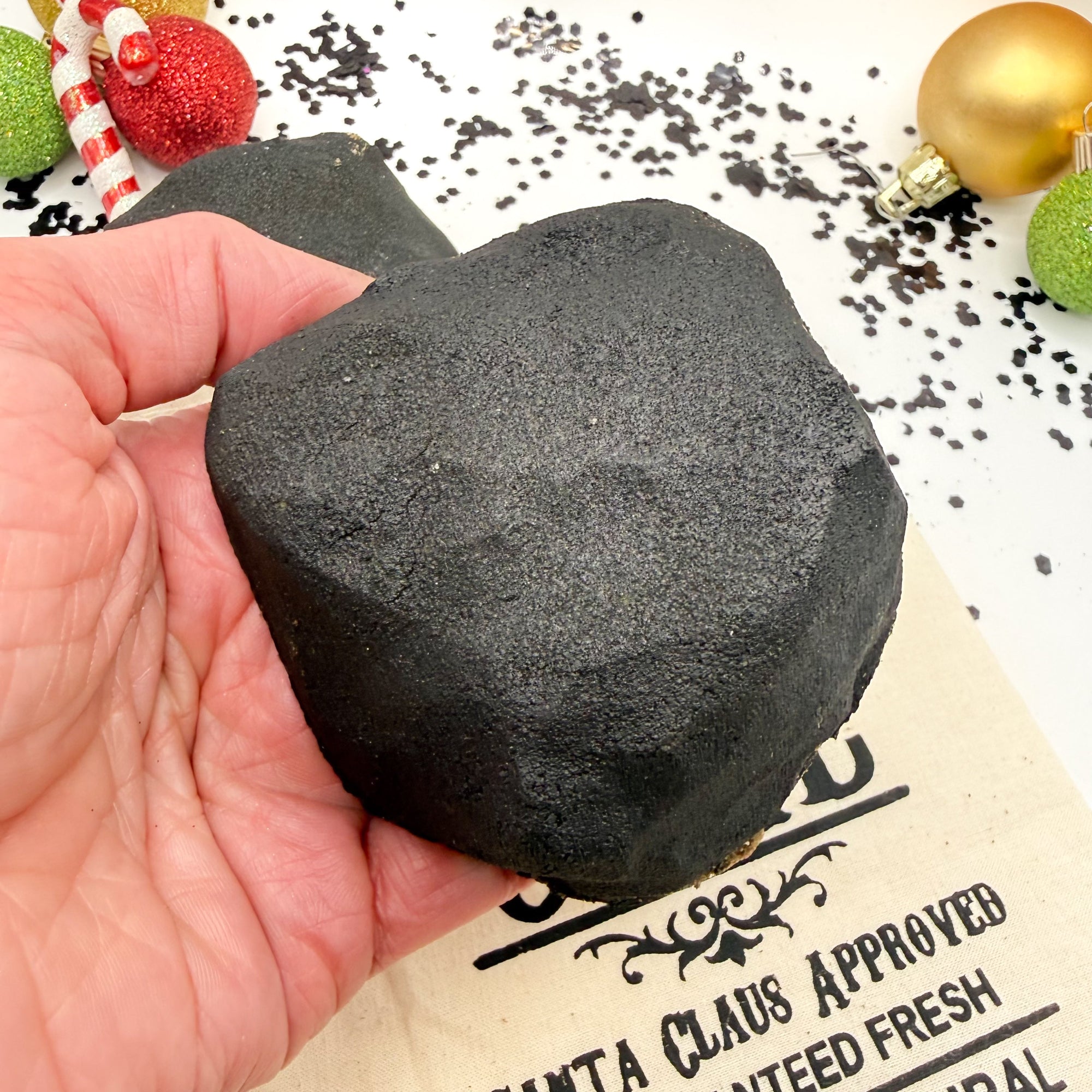 Black lump of coal bath bomb in a hand with Christmas decorations in the background handmade by Rusted Acre Soap Co.
