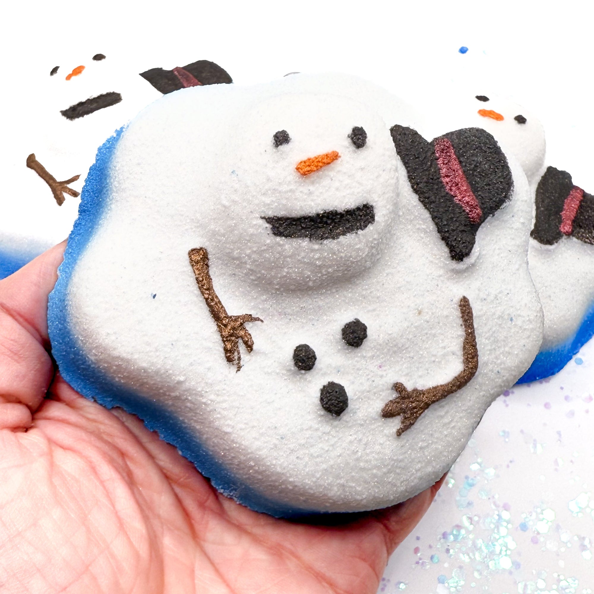 Melting snowman bath bomb held in a hand with a sparkly background hand painted by Rusted Acre Soap Co.
