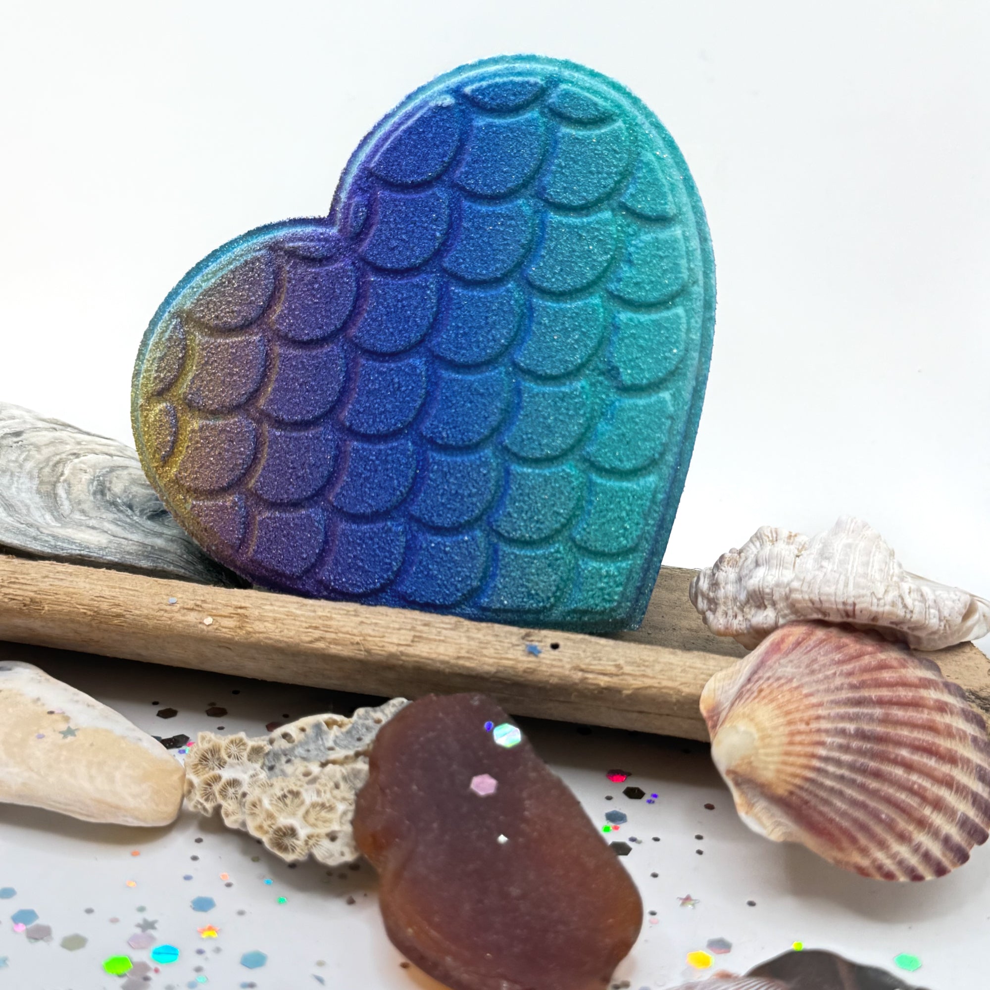 Heart shaped bath bomb with mermaid scale pattern on a white background with seashells and driftwood.
