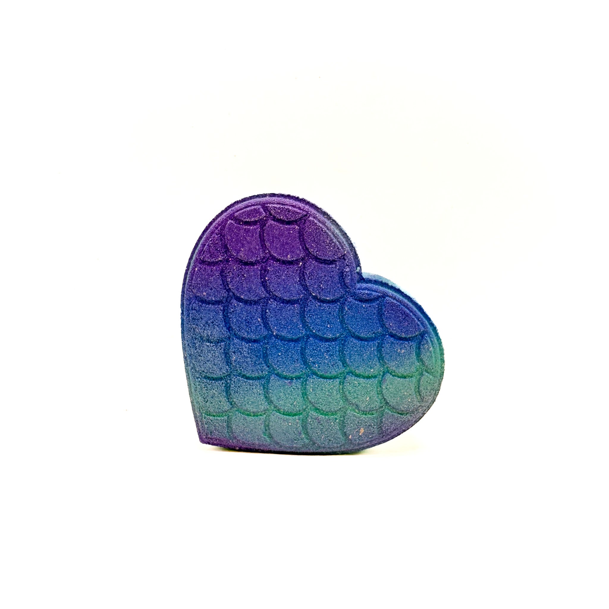 Teal, heart shaped bath bomb with mermaid scales. Purple to teal airbrushed gradient. Gold accent on the left side. Displayed on a rustic wooden background.