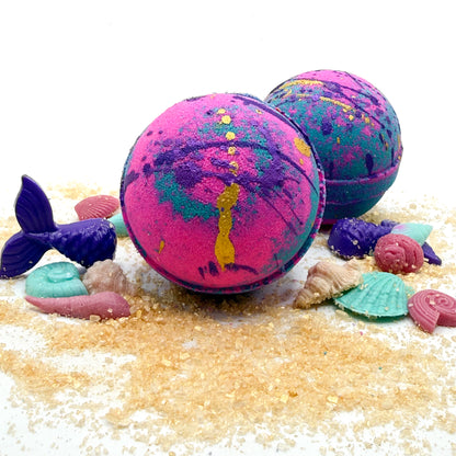 Pink and teal bath bombs scented in mermaid kiss displayed on a sandy surface with sea-themed decorations.