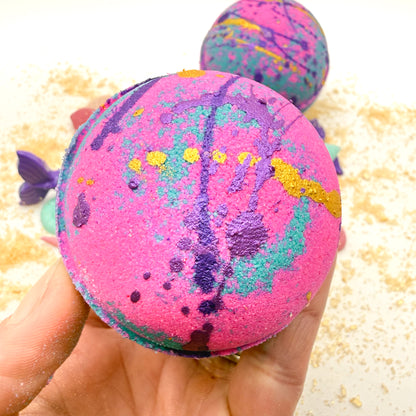 Pink and teal bath bomb held in a hand with a blurred beach themed background.