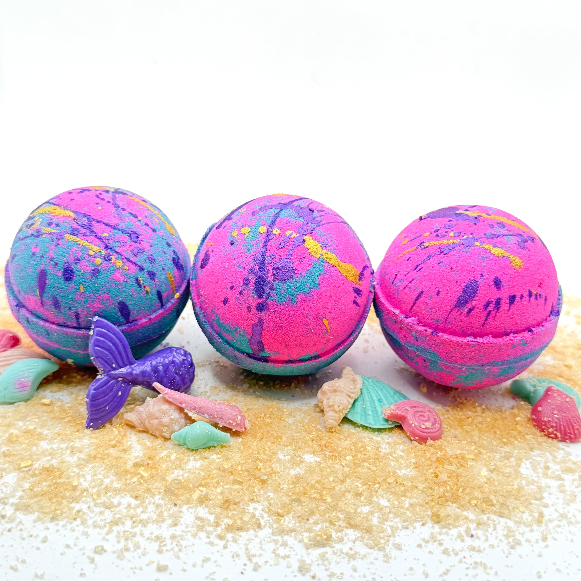 Three pink and teal round mermaid themed bath bombs with a mermaid-themed decorations on a beach setting.
