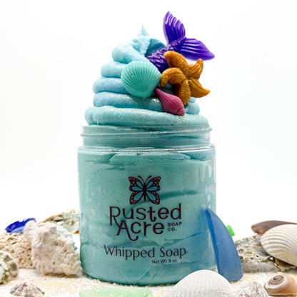 Jar of blue whipped soap from Rusted Acre Soap Co with colorful sea-themed soap embeds surrounded with seashells.