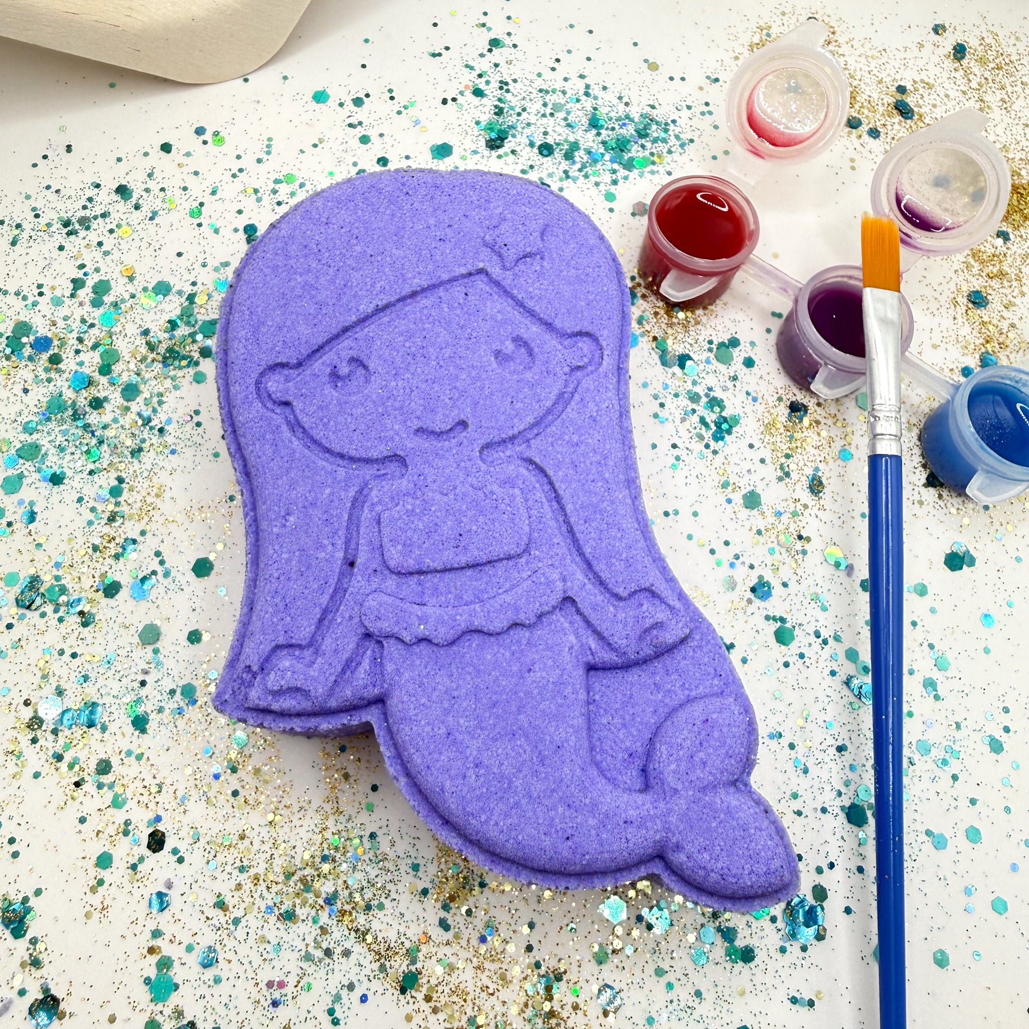 Rusted Acre Soap CO’s purple paint your own mermaid bath bombs on a glittery surface with paint containers and a brush.