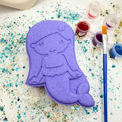 Rusted Acre Soap CO’s purple paint your own mermaid bath bombs on a glittery surface with paint containers and a brush.