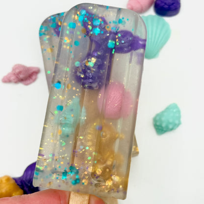 Two hand poured Popsicle soaps with mermaid themed soap embeds and sparkling glitter. 