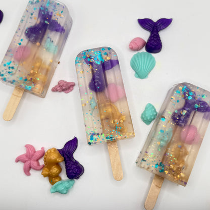 Three popsicle soaps with mermaid themed embeds on a white background with other mermaid soap s and embeds. 