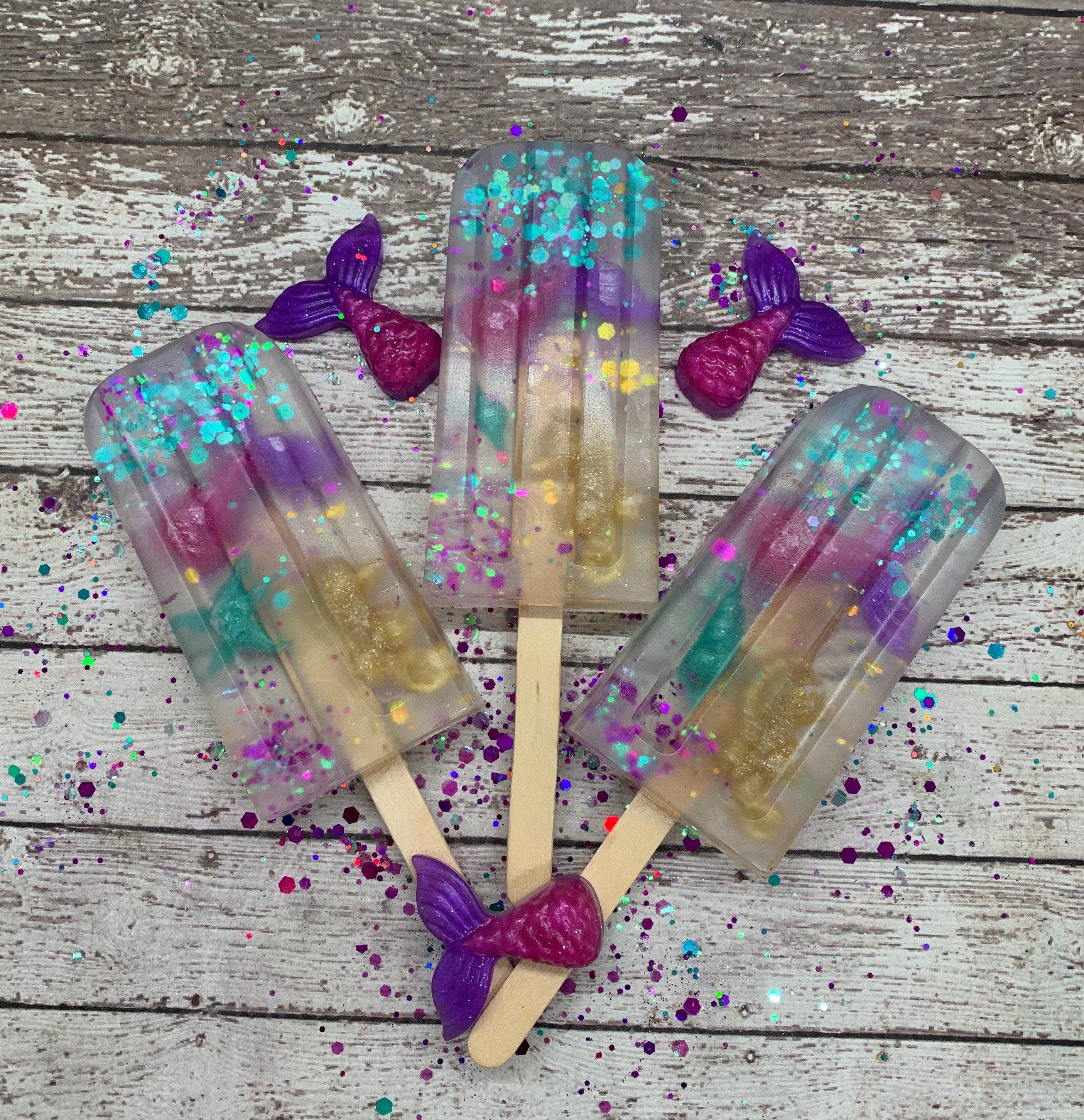 Three mermaid themed clear soaps on a popsicle stick. Embedded in the soap are pink to purple mermaid tail soap and teal sprinkles. 