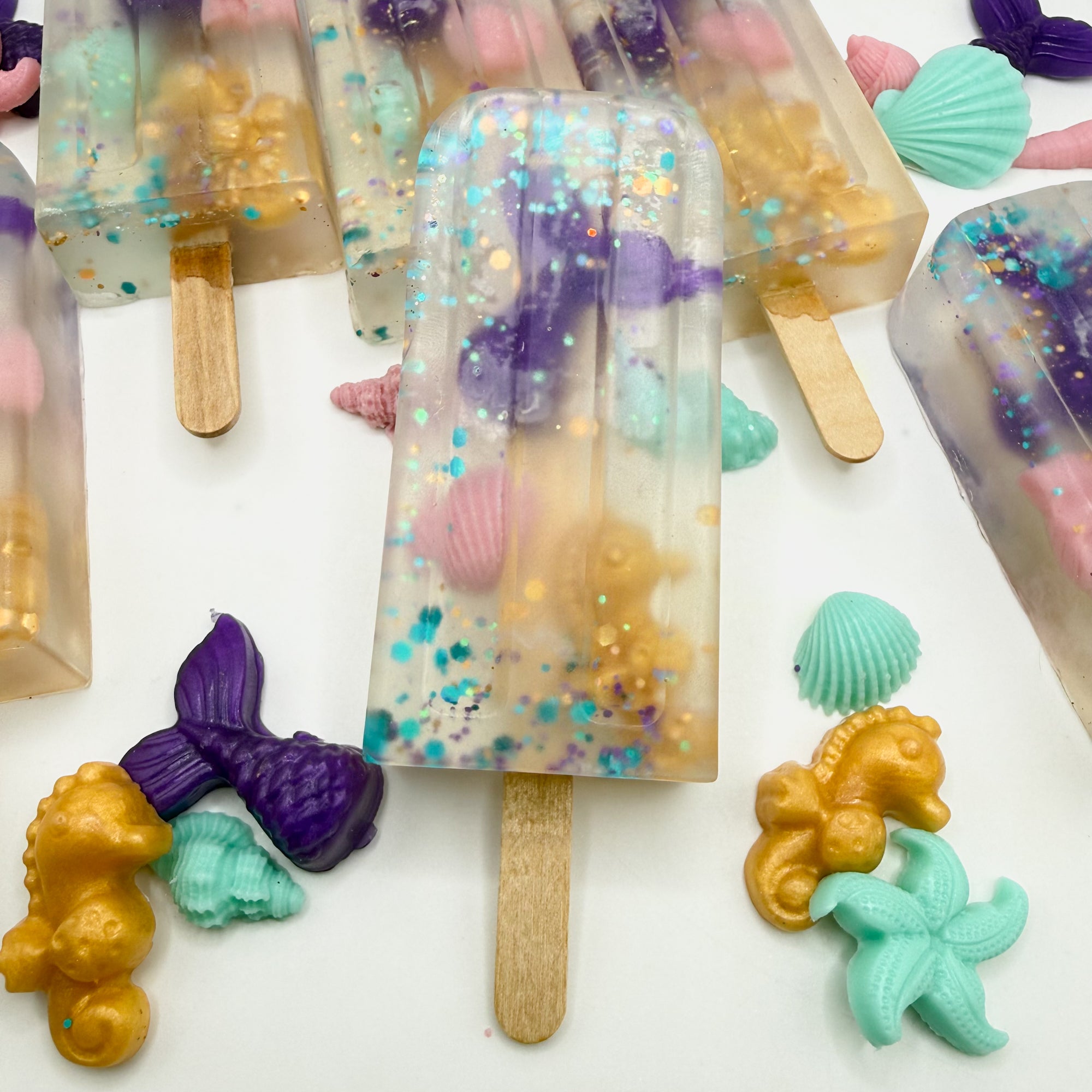 Mermaid themed popsicle soaps on a white background with mermaid themed soap embeds for props and main focus on one popsicle soaps with other soaps in the background. 
