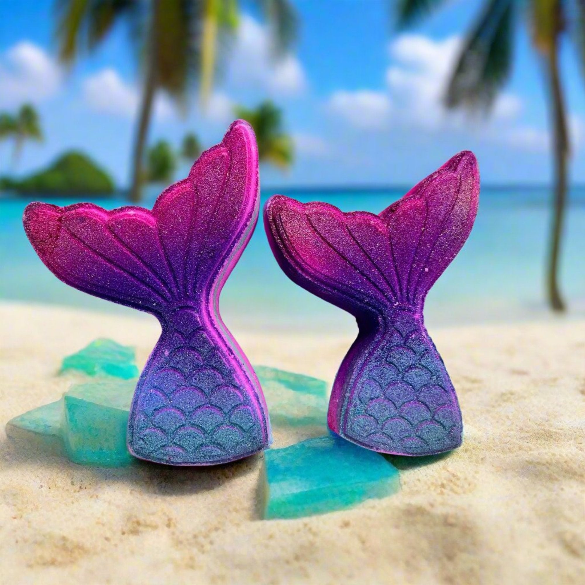 Two mermaid tail shaped bath bombs. Airbrushed in gradient from blue to pink. Displayed on a tropical beach background.