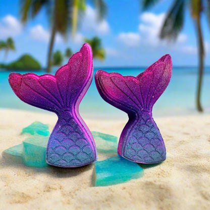 Two mermaid tail shaped bath bombs. Airbrushed in gradient from blue to pink. Displayed on a tropical beach background.