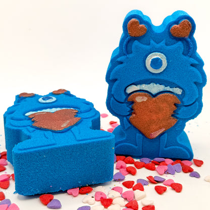 Two blue monster shaped bath bombs. One standing, one laying. Both are holding big red hearts and have red antennae they are hand-painted.