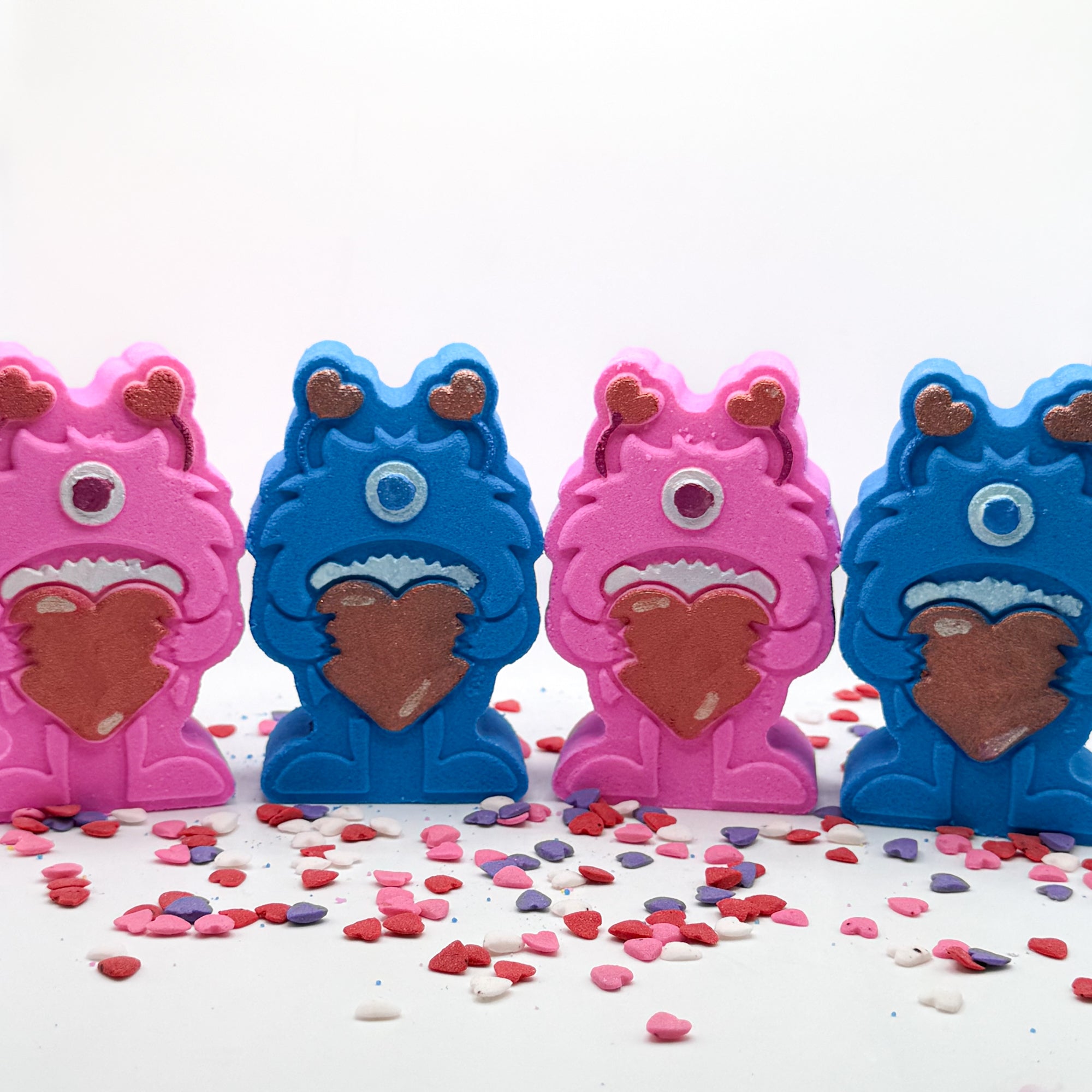 Four monster shaped bath bombs. Two blue, two pink. They are holding big red hearts and have red antennae they are hand-painted