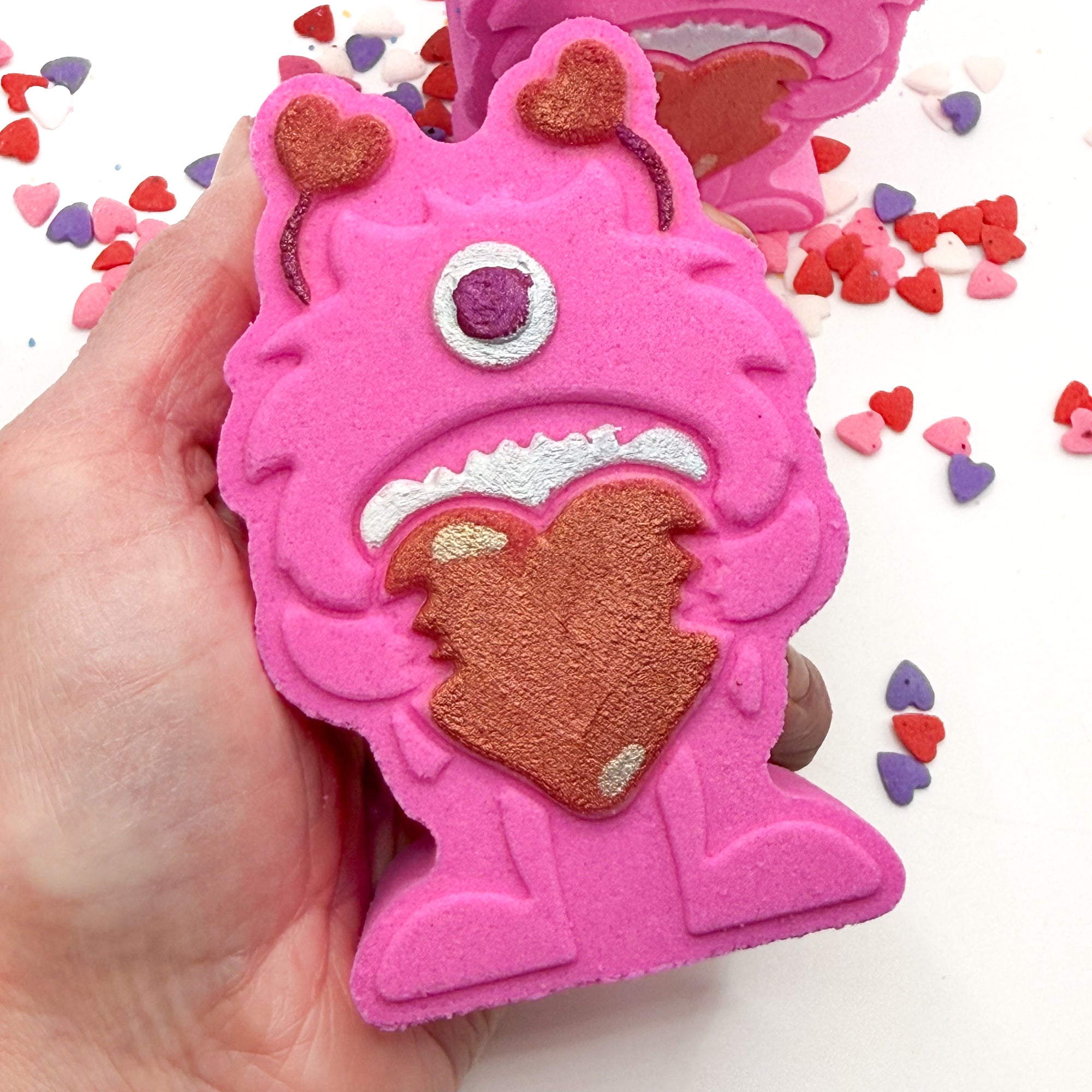 Hand painted Pink monster shaped bath bomb being held in a hand with heart shape candies around it. 
