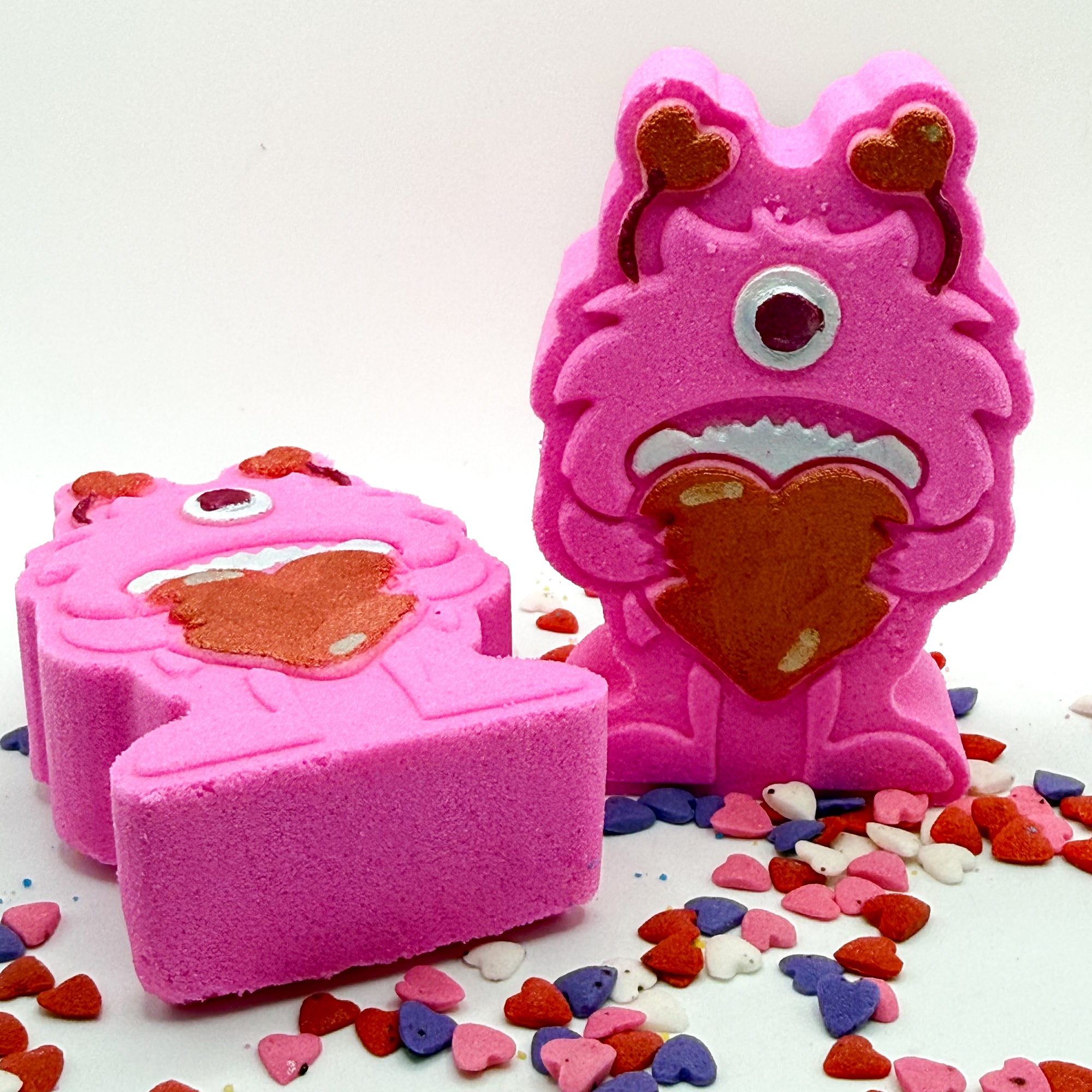 Two pink monster shaped bath bombs. One standing, one laying. Both are holding big red hearts and have red antennae they are hand-painted.