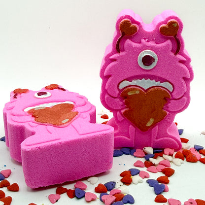 Two pink monster shaped bath bombs. One standing, one laying. Both are holding big red hearts and have red antennae they are hand-painted.