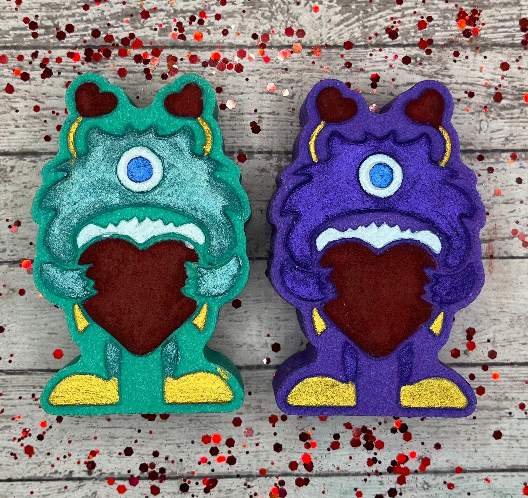 Two monster shaped bath bombs. One teal, one purple. Both are holding big red hearts and have red antennae, one blue eye and gold shoes that are hand-painted.