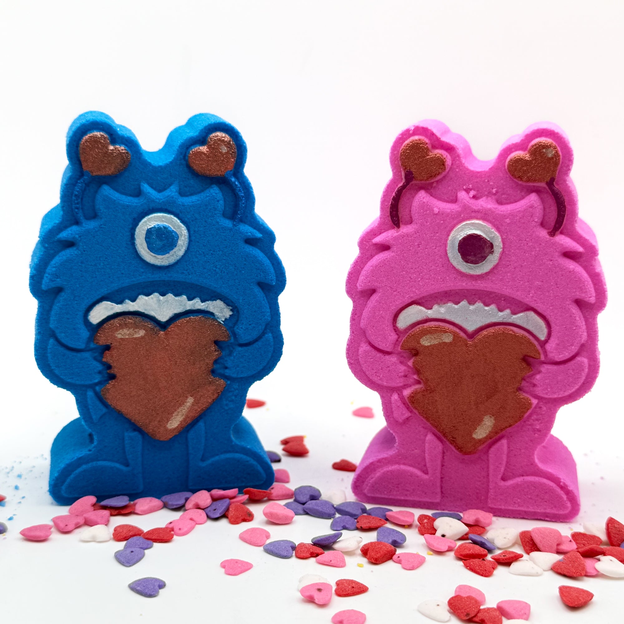 Two hand painted monster shaped bath bombs. One blue, one pink. Both are holding big red hearts and have red antennae.