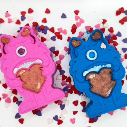 Two monster shaped bath bombs. One blue, one pink. Both are holding big red hearts and have red antennae they are hand-painted.