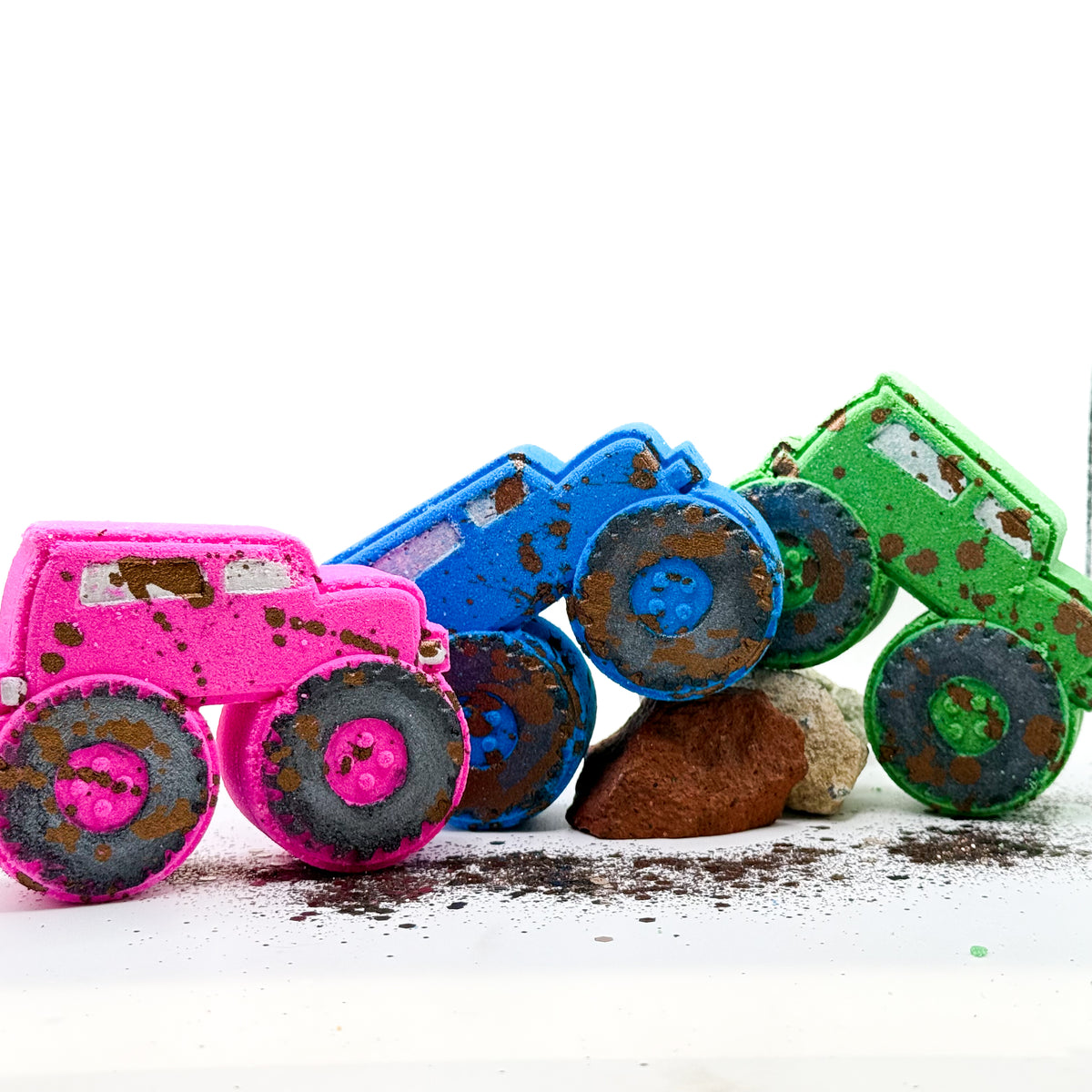 Three colorful monster truck shaped bath bombs in pink, blue, and green, styled with fake mud and rocks on a white background.