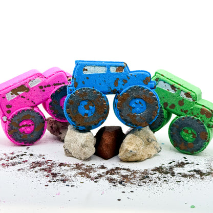 Three monster truck shaped bath bombs in pink, blue, and green, decorated with brown paint to resemble mud and resting on rocks on a white background.
