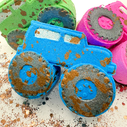 A close up of colorful, muddy truck shaped bath bombs on a white surface. The main subject is a bright blue truck with black and brown glittery mud-splattered details, with other green and pink trucks partially visible in the background.