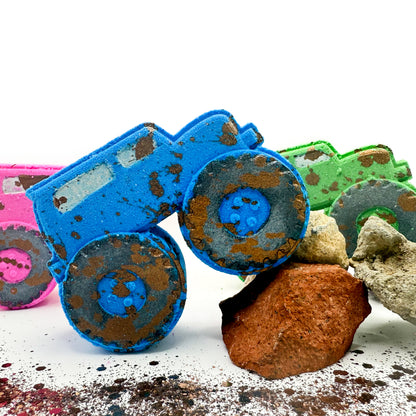 Three monster truck shaped bath bombs in blue, green, and pink,with hand painted mud splatter and placed on rocks on a white surface against a white background.