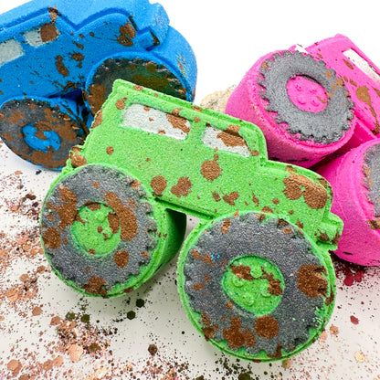 Three truck shaped bath bombs in blue, green, and pink, decorated with glittery black wheels and brown glitter mud splatters, arranged on a white surface.
