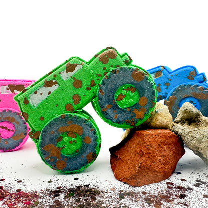 Three monster truck shaped bath bombs in green, blue, and pink, pictured with textured rocks and powder on a white background.