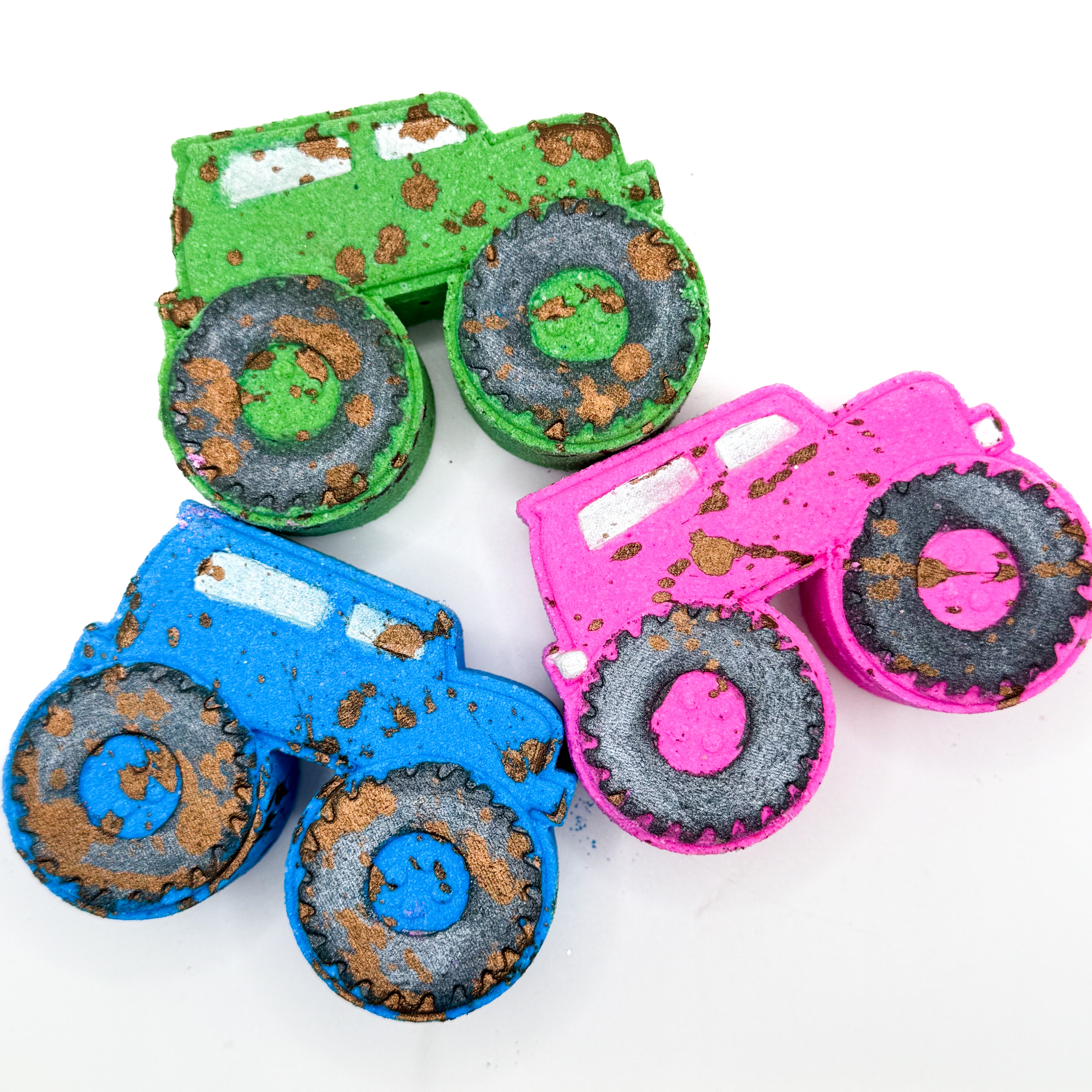 Three monster truck shaped bath bombs in green, blue, and pink, each with  hand painted treaded wheels and brown mud splatter arranged on a plain white background.
