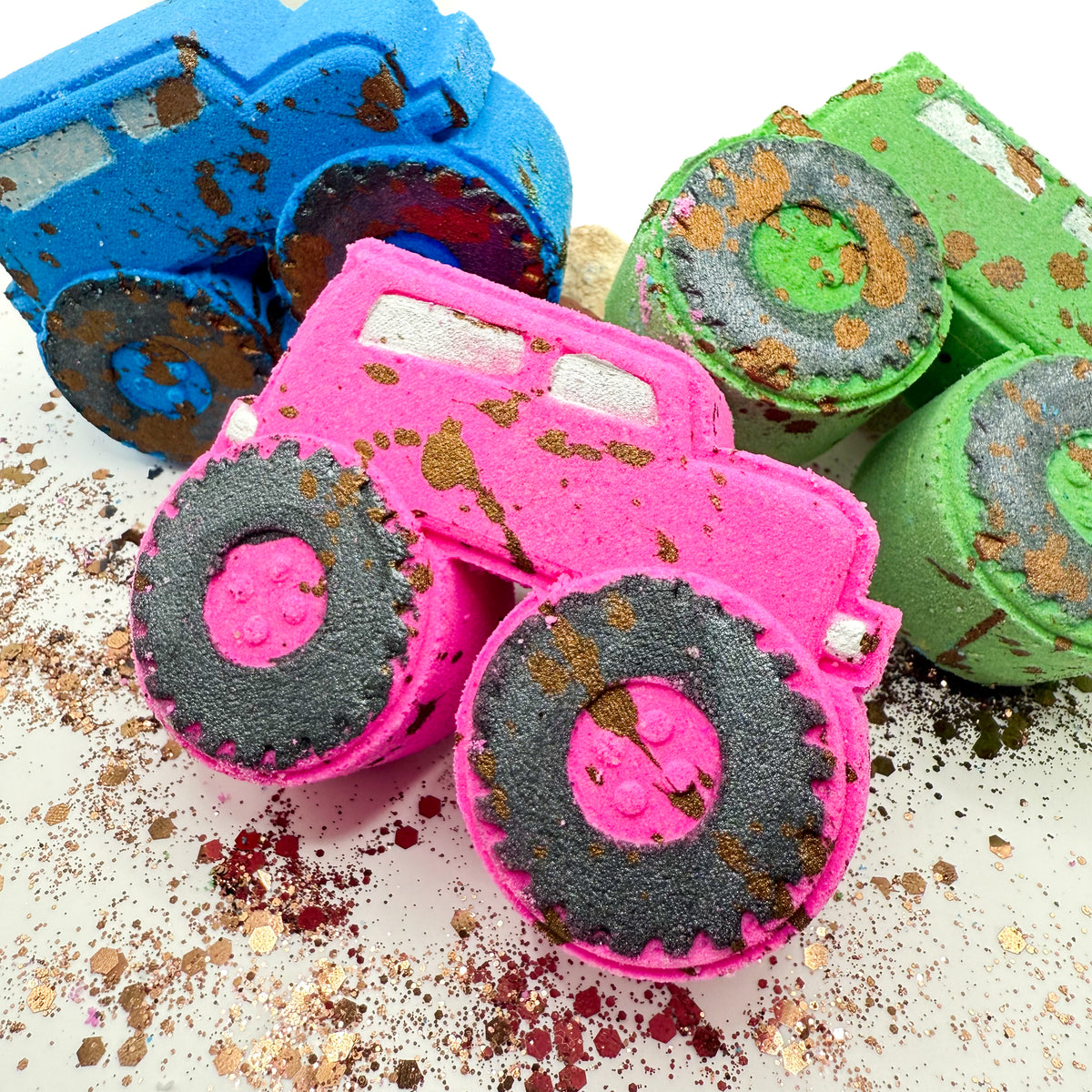 Three colorful monster truck shaped bath bombs or soaps arranged on a white surface with glitter. The front truck is bright pink with black treaded tires and brown splatter, while the other two are blue and green with similar treaded details.
