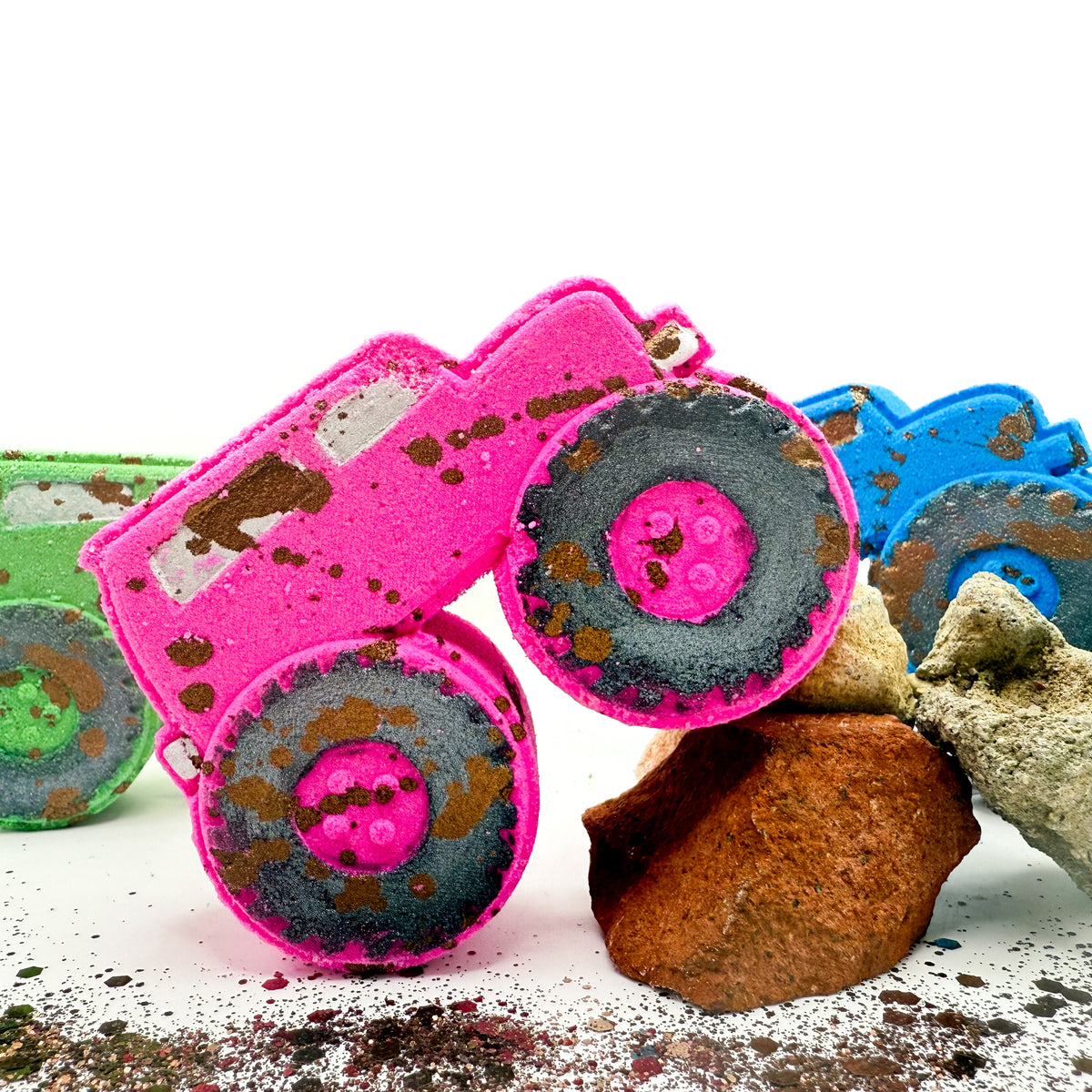 A bright pink monster truck with hand painted black wheels and brown mud like speckling, posed on white with rocks and other colorful bath bombs partially visible in the background.