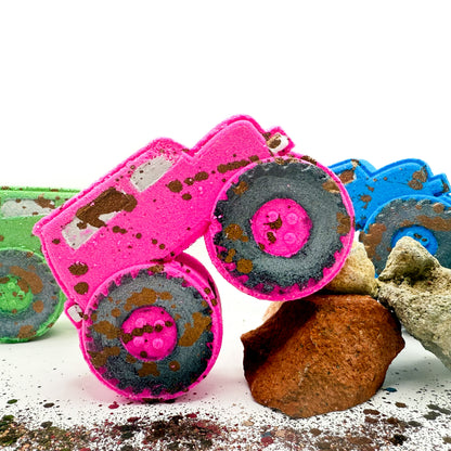 A bright pink monster truck with hand painted black wheels and brown mud like speckling, posed on white with rocks and other colorful bath bombs partially visible in the background.