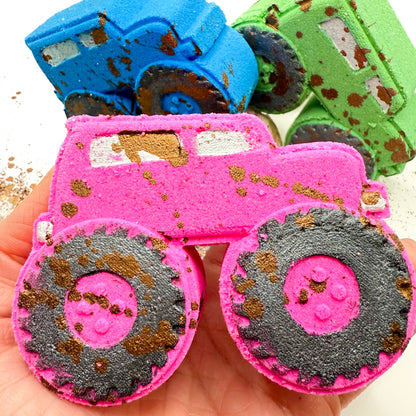 A close up of a hand holding a pink monster truck bath bomb  with hand painted brown mud-splattered and black tires. Two more monster truck bath bombs, one blue and one green, are visible in the background on a white surface.