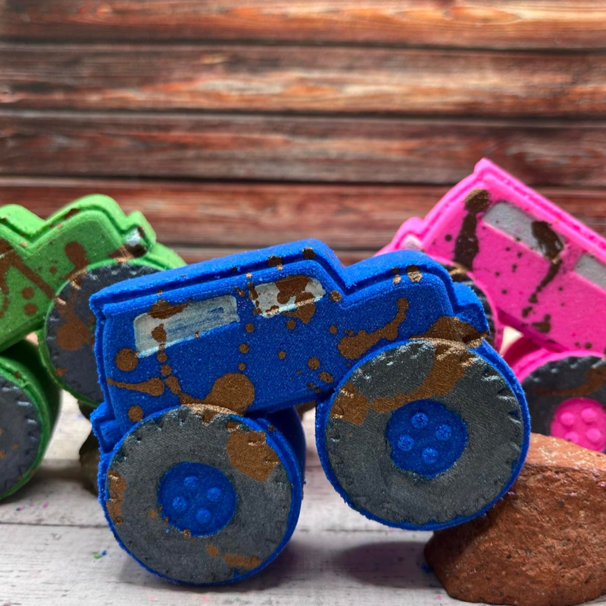 A blue monster truck-shaped bath bomb with hand-painted black wheels, muddy brown splashes, hand painted to look like it just raced through the dirt. This fun, kid-friendly bath bomb is placed on a clean background.