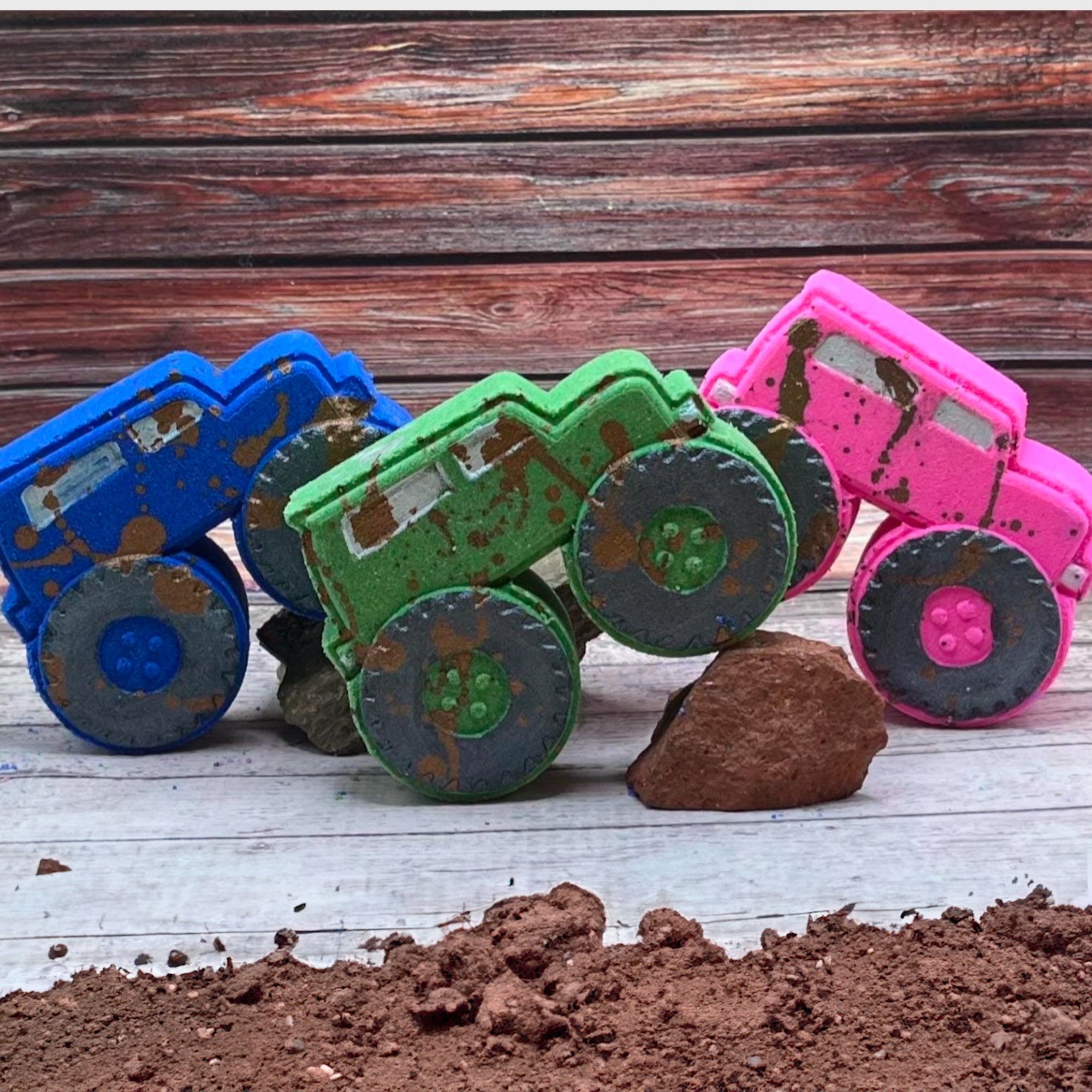 A group of three muddy monster truck bath bombs in pink, blue, and green, each with black wheels and muddy brown splashes, displayed together.