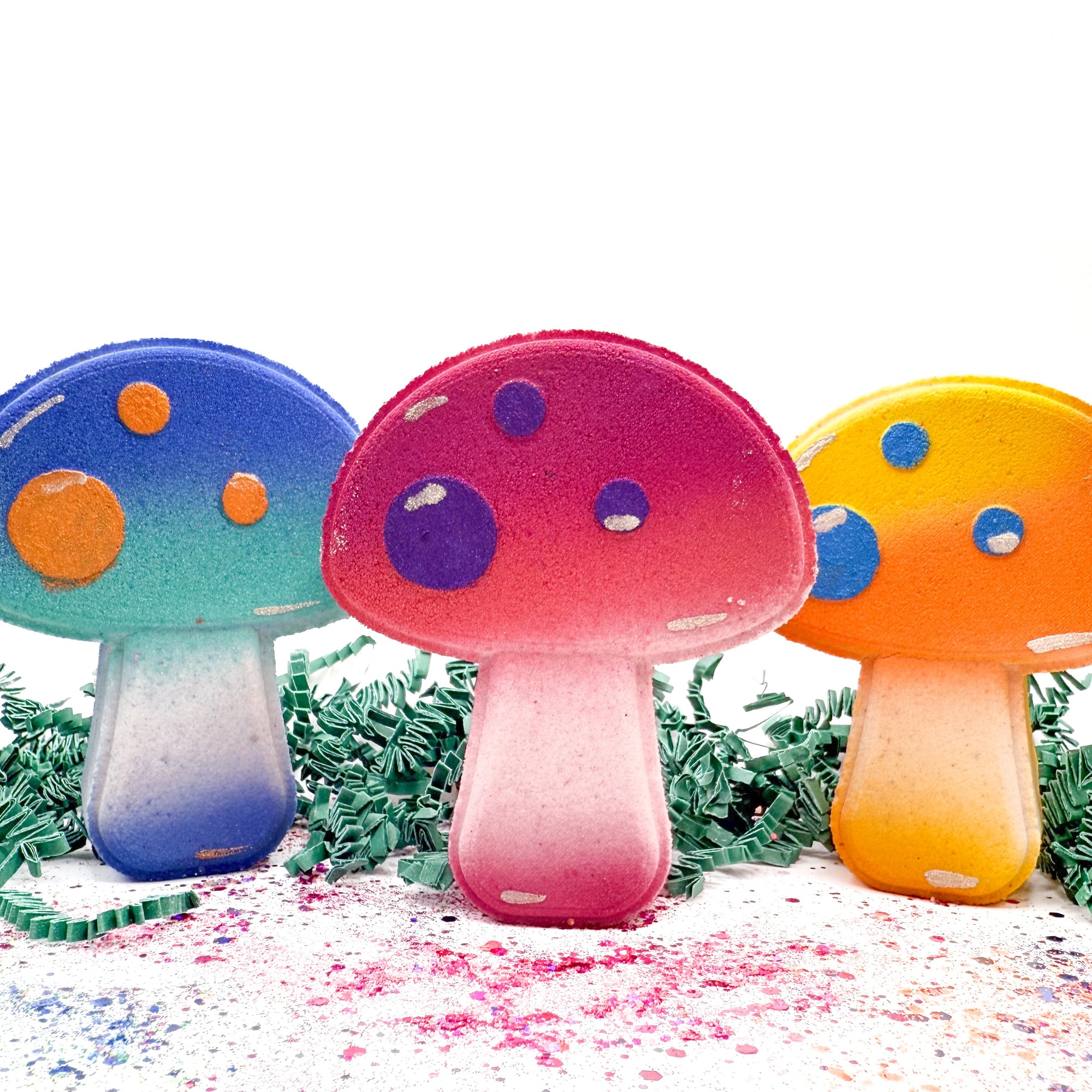 Three colorful mushroom-shaped bath bombs on a white background with greenery.
