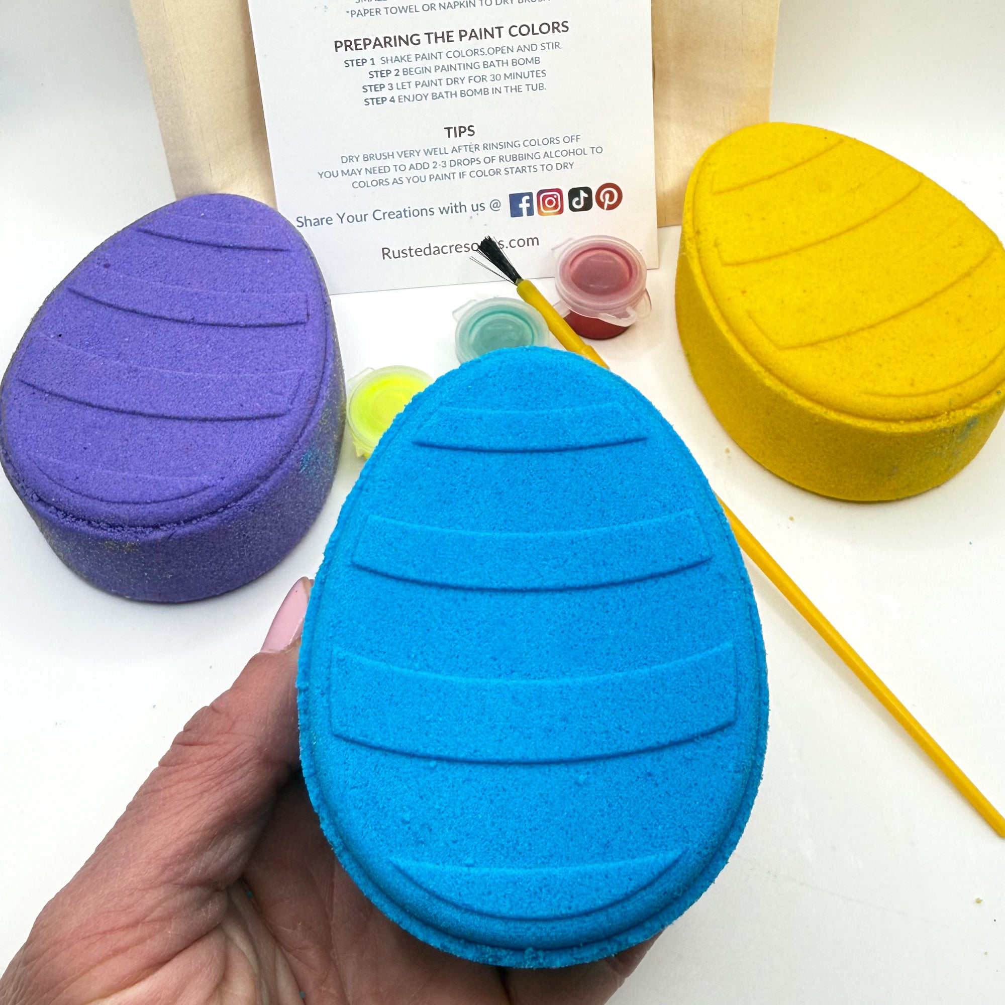 Blue Easter Egg bath bomb held in hand with paint your own kits with paint containers and a brush behind on a white background made by Rusted Acre Soap Co.