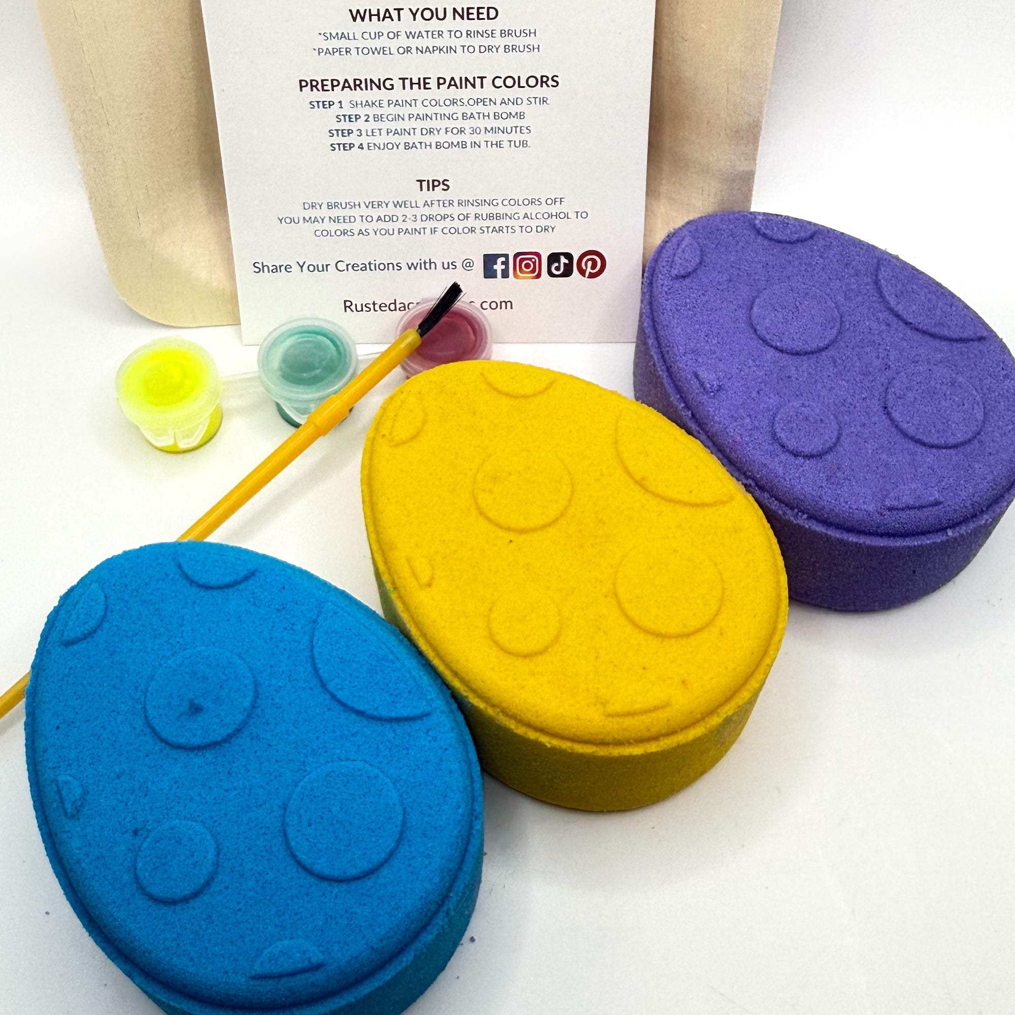 Three Easter Egg bath bombs yellow, blue and purple with paint your own kits and a brush on a white background hand made by Rusted Acre Soap Co.