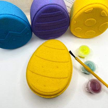 One yellow Easter Egg bath bomb kit with paint containers and a brush on a white background made by Rusted Acre Soap Co with a blue, purple and yellow egg bath bomb in the background. 