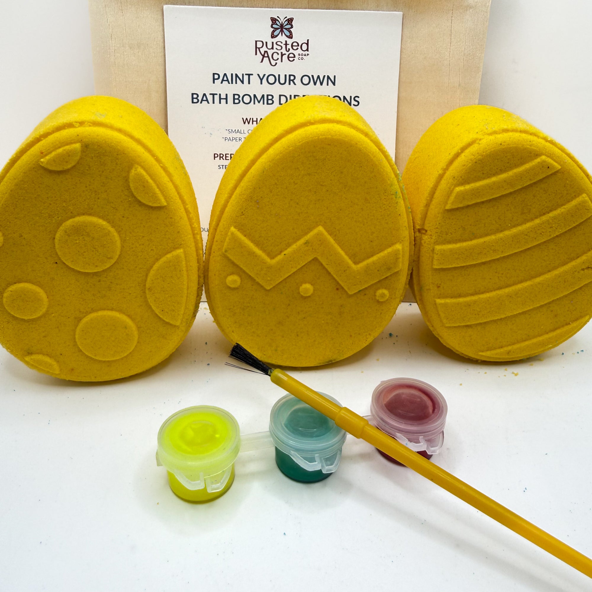 Three yellow Easter Egg bath bomb kits with paint containers and a brush on a white background made by Rusted Acre Soap Co.