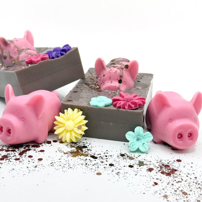 Top and side view of two hand made decorative soaps with pink pig and flowers in a brown mud looking soap. 