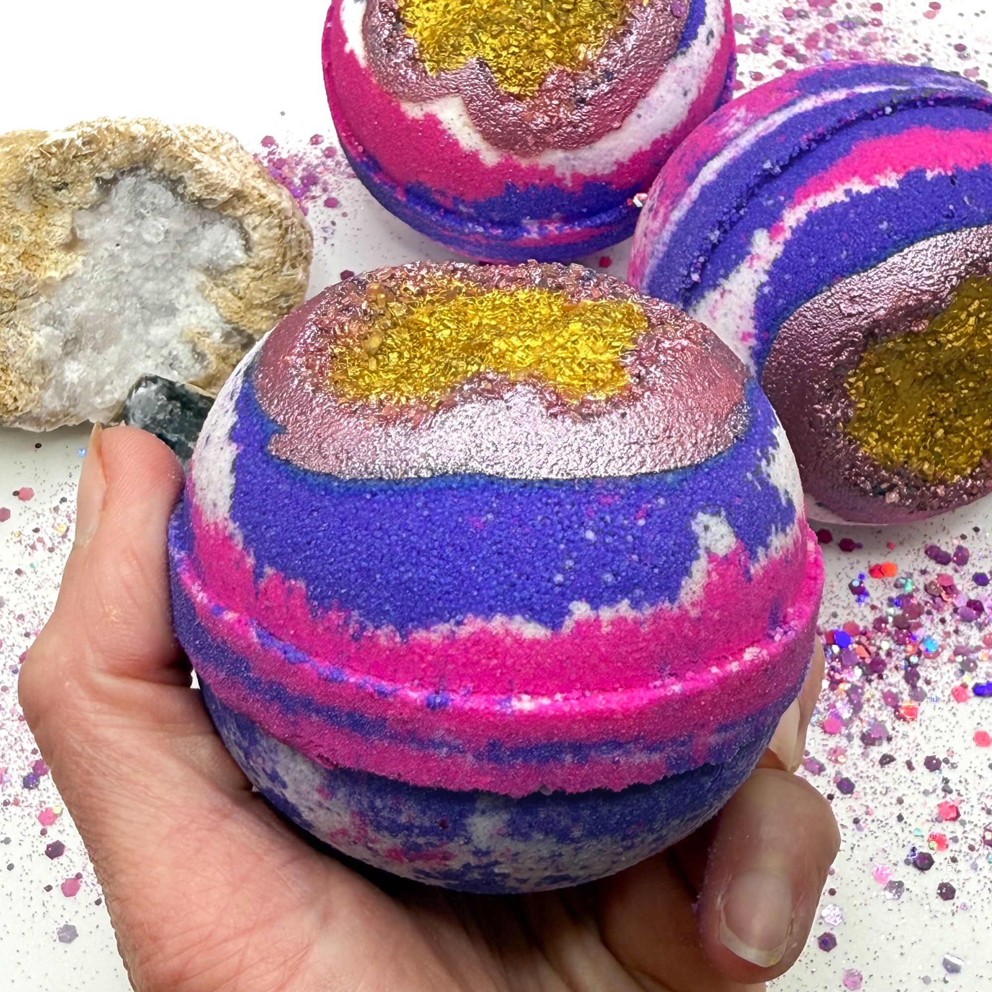 One pink, white and purple bath bomb held in a hand with a gold center resembling a geode on a glittery surface with crystals