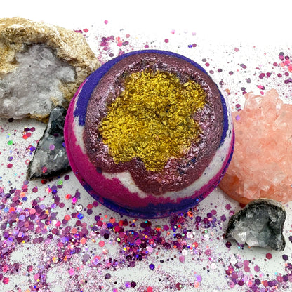 Geode inspired bath bomb pink, purple and white with gold center on a decorative surface with crystals and glitter