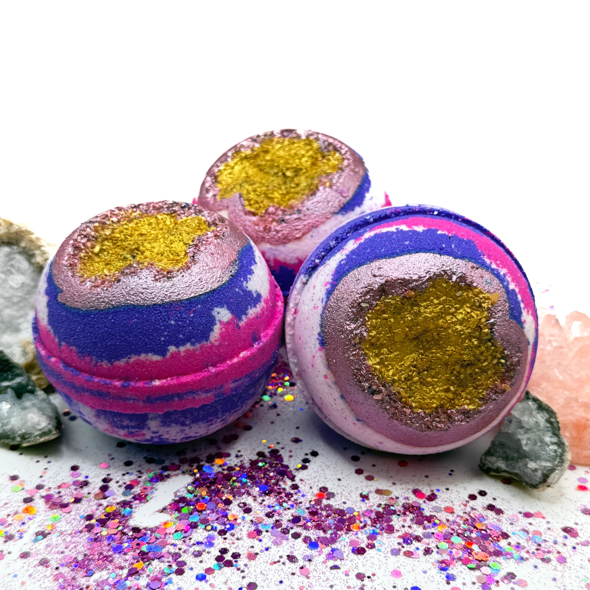 Three pink, white and purple bath bombs with purple and gold centers resembling a geode on a glittery surface with crystals surrounding the bath bombs.