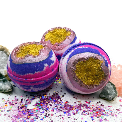 Three pink, white and purple bath bombs with purple and gold centers resembling a geode on a glittery surface with crystals surrounding the bath bombs.