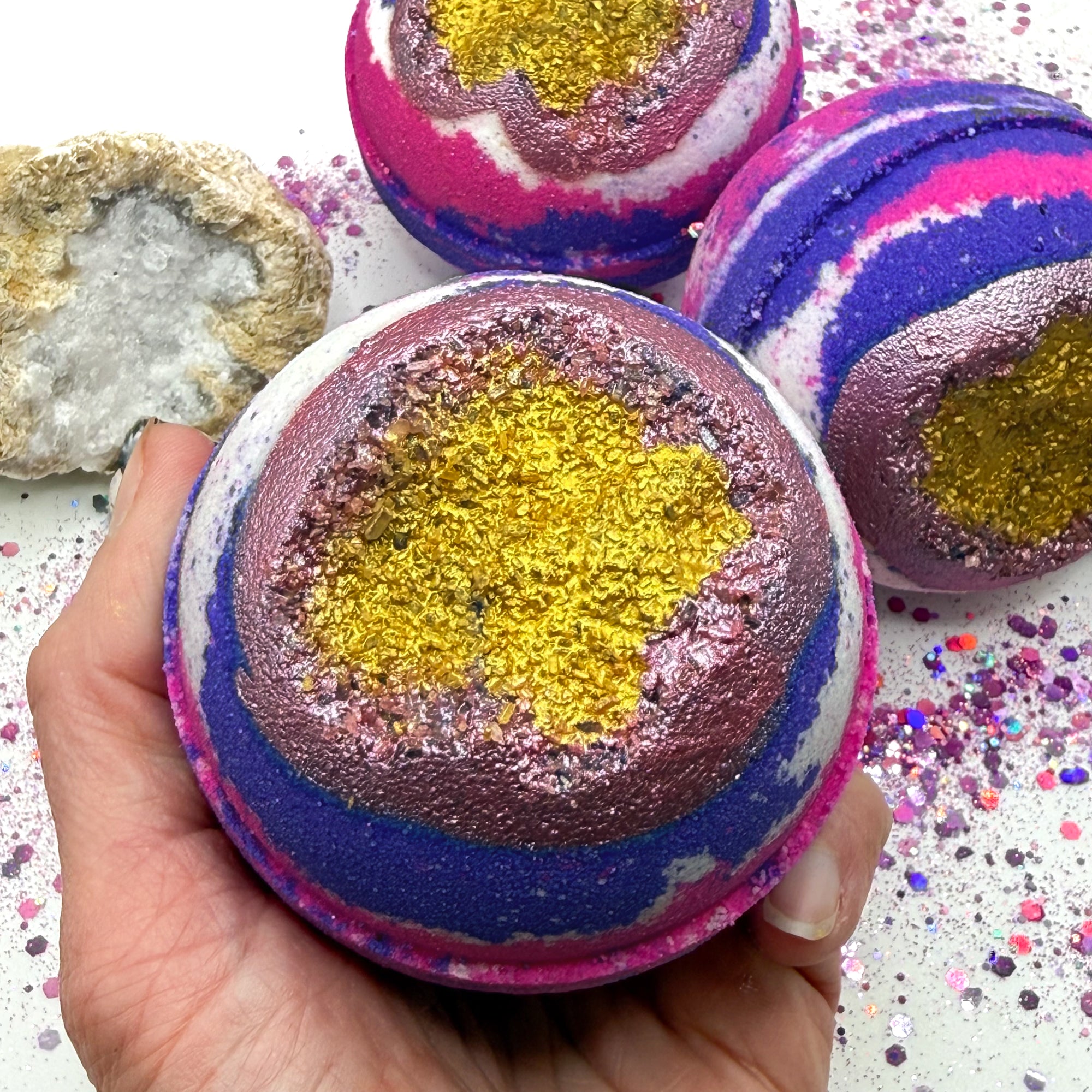 Pink, purple and white bath bomb with geode inspired center held by a hand, surrounded by more bath bombs and decorative elements.