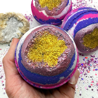 Pink, purple and white bath bomb with geode inspired center held by a hand, surrounded by more bath bombs and decorative elements.